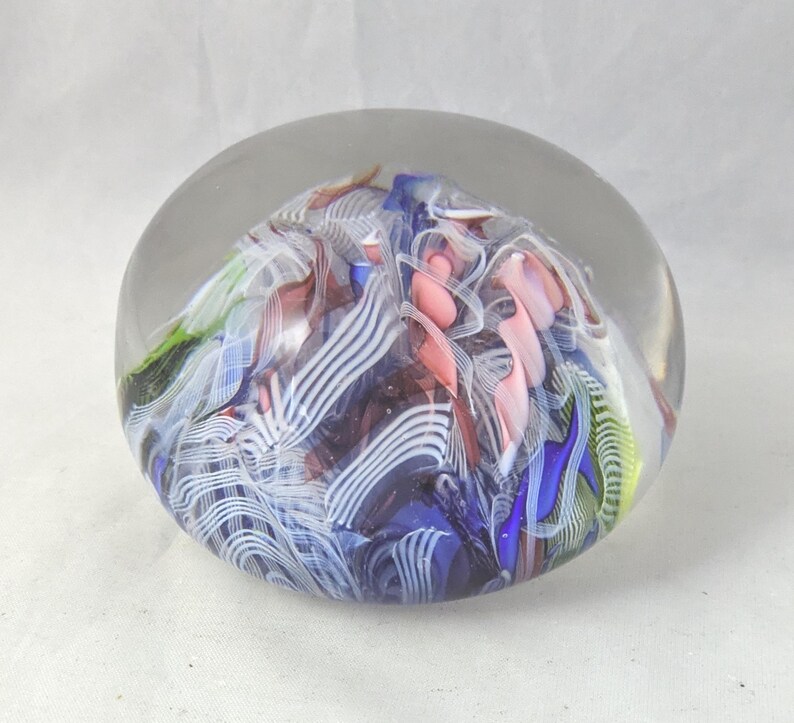 May include: A clear glass paperweight with a rounded dome shape containing an internal design of swirling ribbons in shades of blue, white, pink, and green. The interior features intricate patterns of opaque and translucent glass filaments.