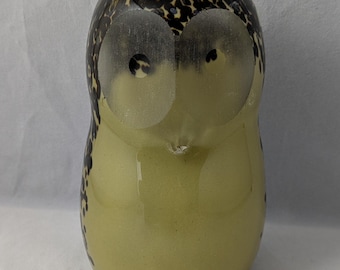 May include: A glass owl figurine with a brown and black spotted pattern. The owl has large, clear eyes and a light yellow body.