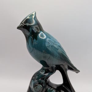 Puede incluir: Una figura de pájaro de cerámica posada sobre una rama, con un acabado brillante en tonos mezclados de azul verdoso profundo, azul oscuro y negro. La escultura representa un ave con una cresta puntiaguda, líneas curvas suaves y una superficie.