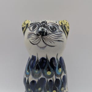 May include: A ceramic cat figurine featuring a cream-coloured head with black painted facial features, whiskers, and yellow-spotted ears. The body is decorated with a repeating pattern of dark blue, light blue, and yellow teardrop-shaped motifs.