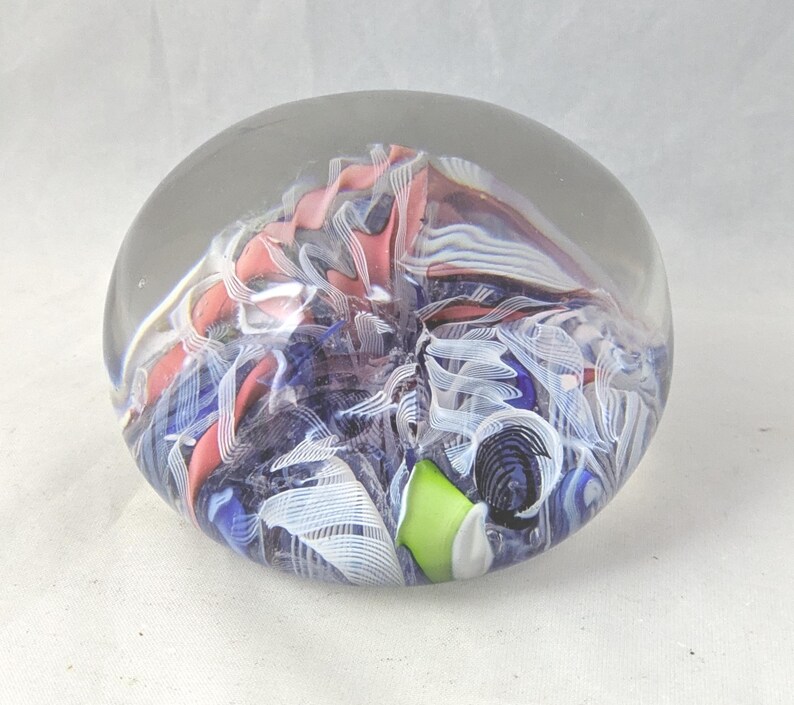 May include: A spherical glass paperweight featuring an internal design of swirling pink, blue, and white ribbons. The clear glass encases a complex pattern of layered colors, including a small bright lime green accent piece near the base.