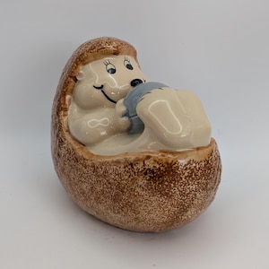 May include: A ceramic hedgehog figurine with a smiling face and a light blue object. The hedgehog is light beige with brown speckles, and the object is held in its arms. The figurine is in a sitting position.