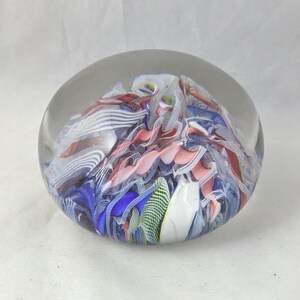 May include: A spherical glass paperweight featuring an internal arrangement of swirling ribbons in shades of blue, pink, white, and green. The clear glass exterior encases the colorful, multi-layered patterns, creating a smooth, rounded object.