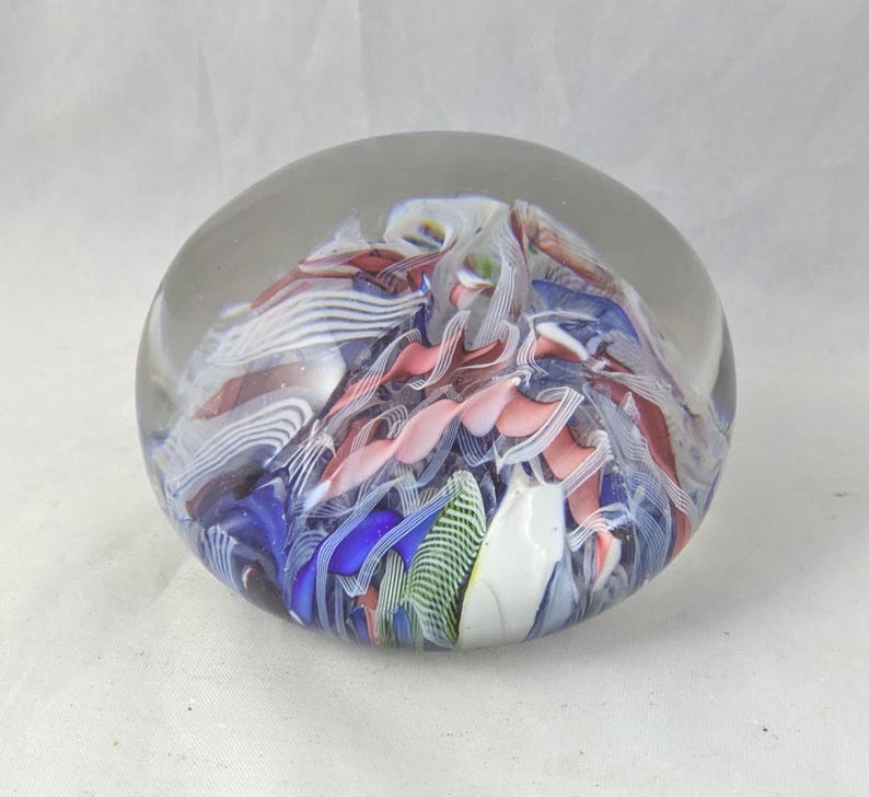 May include: A spherical glass paperweight featuring an internal arrangement of swirling ribbons in shades of blue, pink, white, and green. The clear glass exterior encases the colorful, multi-layered patterns, creating a smooth, rounded object.