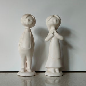 Può includere: Due figurine in ceramica bianca di un ragazzo e una ragazza in piedi uno accanto all'altro. Il ragazzo indossa un abbigliamento semplice e la ragazza indossa un vestito. Entrambe le figurine hanno un design semplice e infantile.