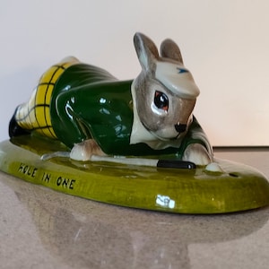Peut inclure: Une figurine en céramique d'un lapin habillé d'une tenue de golf verte et jaune, tenant un club de golf et une balle de golf. La figurine est couchée sur le ventre sur une base ovale verte avec les mots "Hole in One" inscrits dessus.