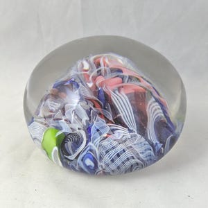 May include: A spherical glass paperweight featuring an internal design of swirling ribbons in shades of blue, white, pink, and a single bright green accent. The clear glass exterior encases the intricate, multi-colored patterns within.