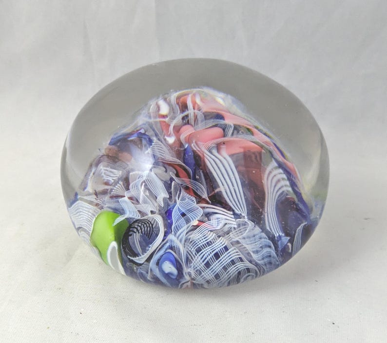 May include: A spherical glass paperweight featuring an internal design of swirling ribbons in shades of blue, white, pink, and a single bright green accent. The clear glass exterior encases the intricate, multi-colored patterns within.
