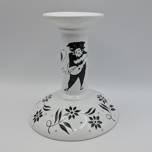 May include: White ceramic candlestick holder with a black illustration of a musician playing a stringed instrument. The base features black floral and leaf designs. The candlestick holder has a wide base and a flared top.