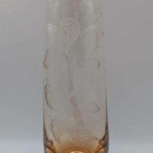 May include: A clear glass vase with an etched leaf design. The vase has a cylindrical shape with a slightly flared rim and a rounded, amber-coloured base. The etched leaves are arranged in a vertical pattern.