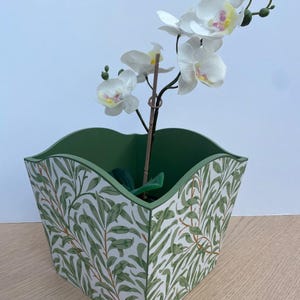 May include: A square green and white patterned planter with a decorative floral design, holding a white orchid with yellow and pink accents. The planter is sitting on a light wood surface.
