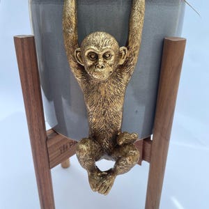 May include: A grey ceramic planter pot held by a gold-coloured monkey figurine that hangs over the rim. The planter is supported by a wooden stand with four legs. The monkey has detailed fur texture and is positioned vertically on the front of the pot.