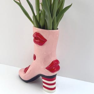 May include: A light pink ceramic boot-shaped vase featuring raised, glossy red lips on the exterior. The heel of the boot is decorated with horizontal red and white stripes, and the vase contains green tulip stems extending from the top opening.