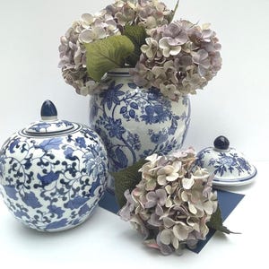 May include: A selection of blue and white porcelain vases and lidded jars with floral designs. A large vase displays a bouquet of lavender hydrangeas. Additional hydrangeas and a small lidded jar are also present.