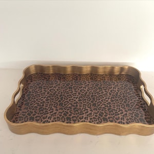 May include: A rectangular serving tray with a wavy, scalloped edge and a metallic gold finish. The interior base of the tray features a brown and black leopard print pattern. The tray includes two integrated handles on the shorter sides.