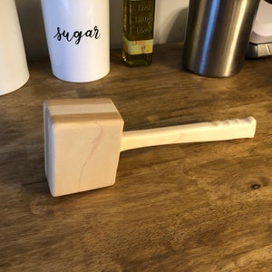 May include: A light-colored wooden mallet with a rectangular head and a long, rounded handle. The handle has a grooved grip for secure handling. The mallet is made of light-colored wood and is designed for striking.