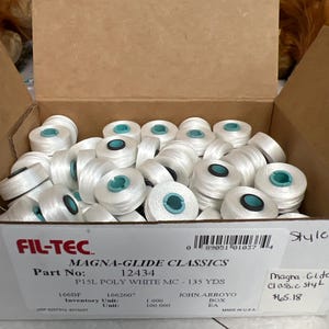 May include: A cardboard box filled with white Magna-Glide Classics polyester bobbins, each featuring a teal plastic center. The box is labeled Fil-Tec, Part No: 12434, P15L Poly White MC - 135 Yds, and includes a barcode and inventory details.