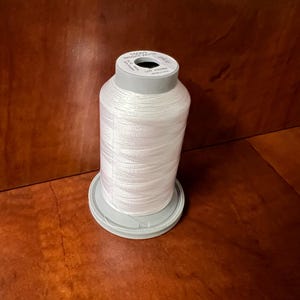 May include: A spool of white thread, labeled "1000S BRIGHT WHITE", sits on a gray plastic base. The thread is tightly wound, and the spool is set against a brown background. The thread is likely used for sewing or embroidery projects.