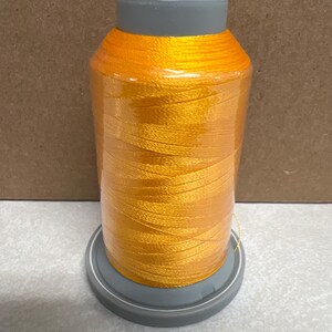 May include: A large spool of vibrant orange embroidery thread wrapped around a gray plastic core. The thread is tightly wound in a cylindrical shape, featuring a smooth, shiny texture. The spool is set on a circular gray base with a matching gray cap.