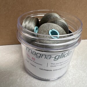 May include: A clear plastic container filled with multiple spools of metallic silver Magna-Glide No. 40 trilobal polyester thread. The label on the container displays the text Magna-Glide, No. 40, Trilobal Polyester, Col. 10401, and German Granit.