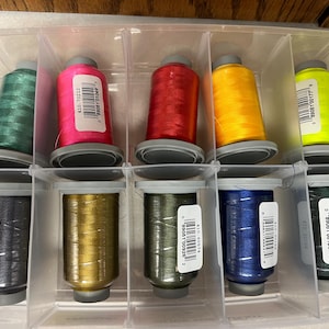 May include: A clear plastic organizer filled with spools of colorful embroidery thread. The spools are in various colors, including green, pink, red, yellow, and blue. Each spool has a gray base and a label with a barcode.