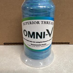 May include: A spool of Superior Threads OMNI-V variegated thread in shades of blue and teal. The white label features the text Superior Threads, OMNI-V, poly-wrapped poly-core variegated thread, and recommended needle sizes #18 for longarm and #90/14 for home.
