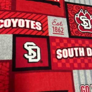 May include: Red and gray patterned fabric featuring the words "South Dakota" and "Coyotes" in white lettering. The fabric also includes the letters "SD" and the year "1862". The design is a mix of squares and stripes.