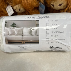 May include: A white plastic-wrapped Cuddle Dreamz pillow form with a label showing a 14 inch by 14 inch size. The packaging features a photograph of white throw pillows on a sofa, a list of available sizes, and text indicating 50% shredded memory foam and 50% gel fiber fill with a 100% polyester cover.