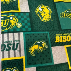 May include: A fleece fabric featuring a patchwork design of North Dakota State University Bison logos and text. The pattern includes yellow bison heads and full-body figures on green backgrounds with various textures, alongside grey stripes and text.
