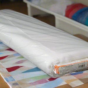 May include: A rectangular, white, packaged item wrapped in clear plastic. The item rests on a colorful, patterned fabric. A bed frame with a colorful blanket is visible in the background.