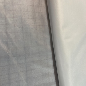 May include: A piece of white fabric featuring a subtle grid pattern on the left side and a smooth, solid white texture on the right side. The material appears lightweight and semi-translucent, showing a clear square weave structure throughout.
