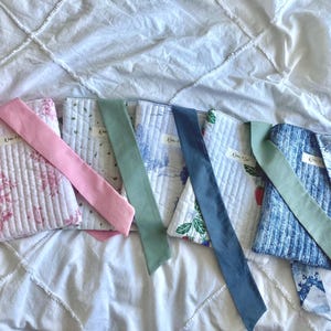 May include: Five rectangular quilted fabric pouches with long fabric straps. The pouches feature various patterns including pink floral, blue toile, and strawberry prints. Each pouch has a small white rectangular label with black text attached.