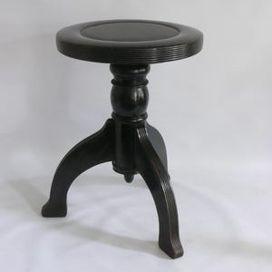 Puede incluir: Un taburete de madera de color marrón oscuro con un asiento circular de superficie lisa y plana con un borde estriado. El taburete se apoya en una base central de pedestal que se divide en tres patas curvas y ranuradas.