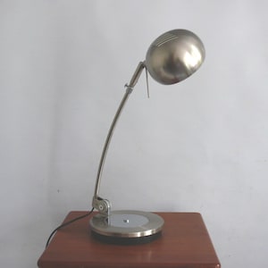 May include: A brushed metal desk lamp featuring a spherical head with linear ventilation slits, a slender adjustable arm, and a circular base. The lamp has a metallic finish and a black power cord extending from the base, showcasing a functional design.
