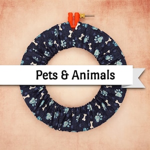 May include: A circular embroidery hoop covered in navy blue fabric with a pattern of light blue paw prints and white bones. The text "Pets & Animals" is displayed on a white banner across the center of the hoop.