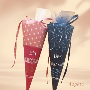 Könnte beinhalten: Zwei personalisierte Schultüten. Eine ist rosa mit weißen Sternen, die andere marineblau mit weißen geometrischen Formen. Beide haben einen roten Boden, den Namen des Schülers, den Schulnamen und das Jahr 2026.