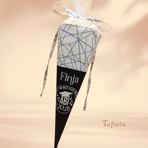 K&ouml;nnte beinhalten: Ein kegelf&ouml;rmiger Geschenkebeutel mit einem schwarz-wei&szlig;en geometrischen Muster. Der untere Teil ist schwarz mit dem Namen "Finja" und "ABI 2026" aufgedruckt. Die Tasche hat ein wei&szlig;es Band und eine dekorative Oberseite.