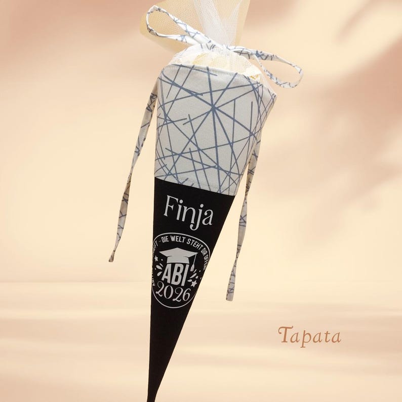 K&ouml;nnte beinhalten: Ein kegelf&ouml;rmiger Geschenkebeutel mit einem schwarz-wei&szlig;en geometrischen Muster. Der untere Teil ist schwarz mit dem Namen "Finja" und "ABI 2026" aufgedruckt. Die Tasche hat ein wei&szlig;es Band und eine dekorative Oberseite.
