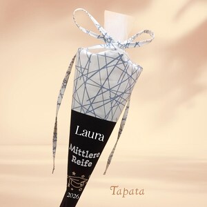 K&ouml;nnte beinhalten: Eine konisch geformte Geschenkt&uuml;te zur Abschlussfeier. Der obere Teil ist wei&szlig; mit einem blauen geometrischen Muster, der untere Teil ist schwarz mit dem Namen "Laura", "Mittlere Reife" und dem Jahr 2026.