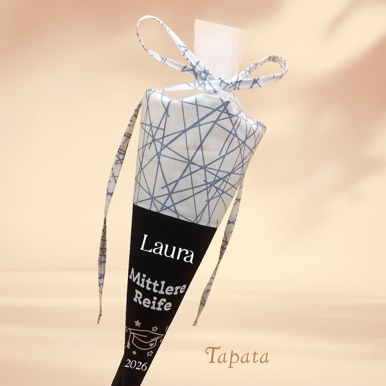 K&ouml;nnte beinhalten: Eine konisch geformte Geschenkt&uuml;te zur Abschlussfeier. Der obere Teil ist wei&szlig; mit einem blauen geometrischen Muster, der untere Teil ist schwarz mit dem Namen "Laura", "Mittlere Reife" und dem Jahr 2026.