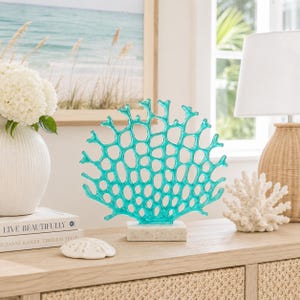 May include: A turquoise coral sculpture on a white marble base. The decorative piece has an open, web-like design. A white coral and a white vase with flowers are also on the wooden surface. A framed beach scene is in the background.