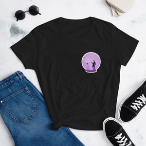 Puede incluir: Una camiseta negra con un gráfico de bola de nieve morada que presenta una pareja bailando dentro. La bola de nieve está sobre un fondo blanco.