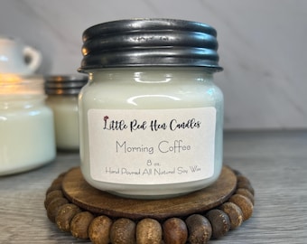 Op de afbeelding: Een potkaars met zilveren deksel, met het opschrift "Morning Coffee" van Little Red Hen Candles. De kaars van 227g staat op een houten onderzetter met een kralenrand. Handgegoten met natuurlijke sojawas.