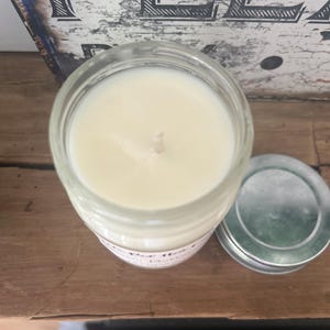 Puede incluir: Una vela en un tarro de vidrio transparente con cera blanca y una sola mecha. La vela est&aacute; sentada sobre una superficie de madera. El tarro tiene una etiqueta blanca con el texto "The Best" y "Love that" en ella.