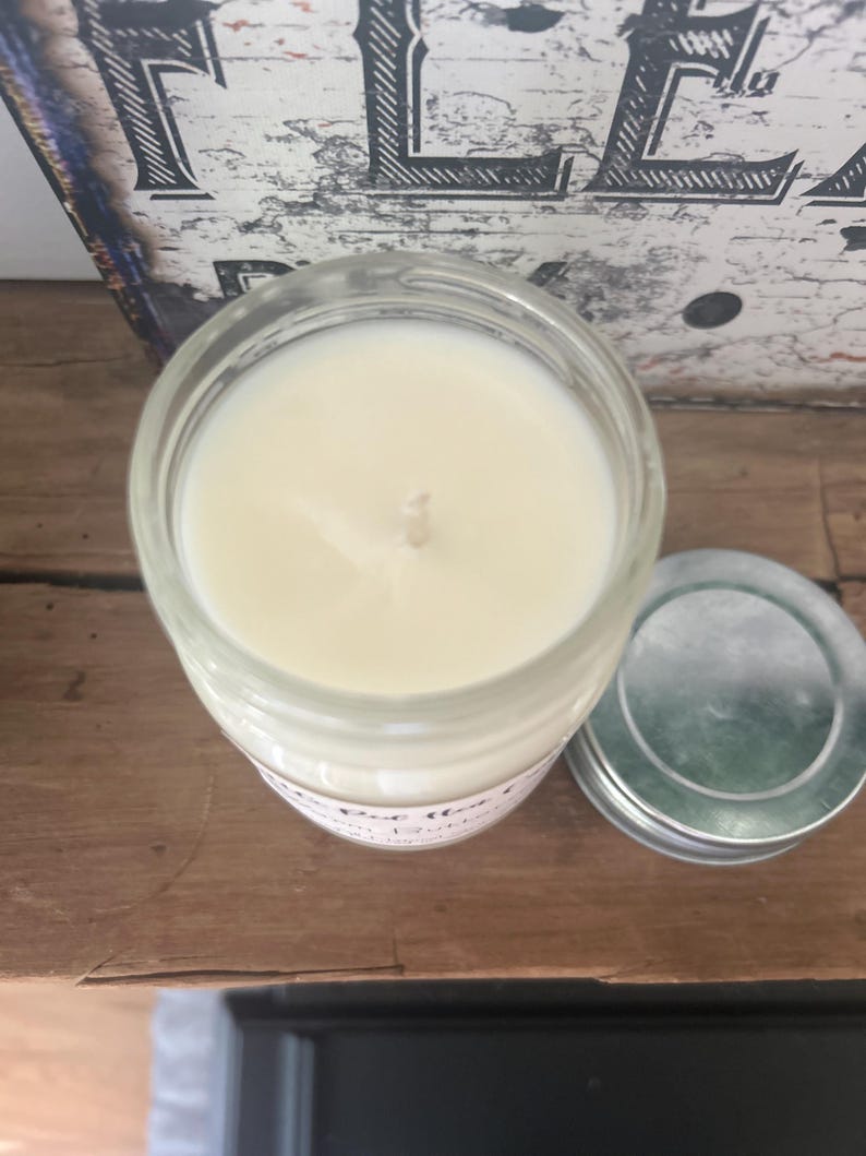 Puede incluir: Una vela en un tarro de vidrio transparente con cera blanca y una sola mecha. La vela est&aacute; sentada sobre una superficie de madera. El tarro tiene una etiqueta blanca con el texto "The Best" y "Love that" en ella.