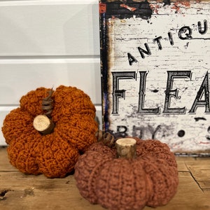 May include: Two crocheted pumpkins, one orange and one brown, with wooden stems. The pumpkins are sitting on a wooden surface in front of a sign that reads "Antique Flea Buy Sell".