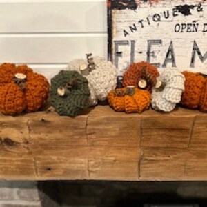 May include: A collection of eight crocheted pumpkins in various colours, including orange, white, and green. The pumpkins are arranged on a wooden mantelpiece.