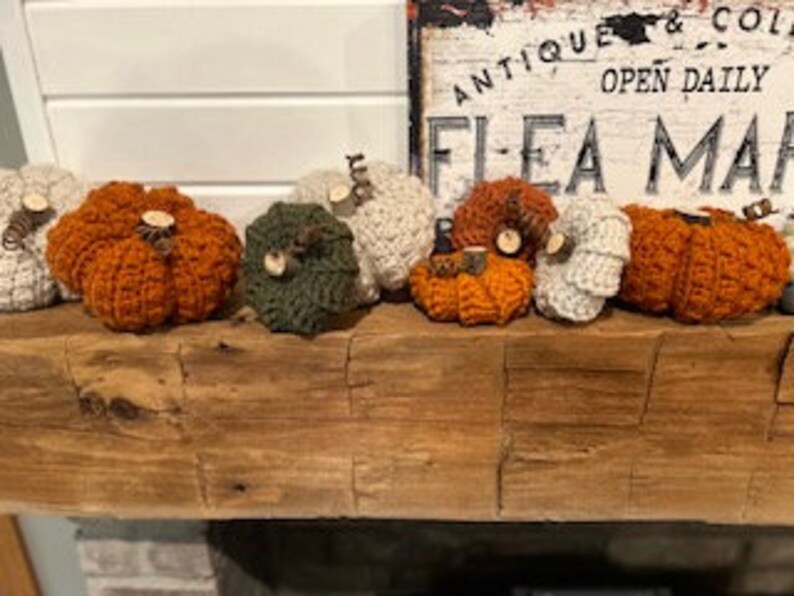 May include: A collection of eight crocheted pumpkins in various colours, including orange, white, and green. The pumpkins are arranged on a wooden mantelpiece.