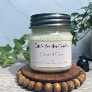 May include: A jar candle with a silver lid, labeled "Caramel Corn" from Little Red Hen Candles. The candle is on a wooden coaster with a beaded edge. The candle is 8 oz and made with all-natural soy wax.