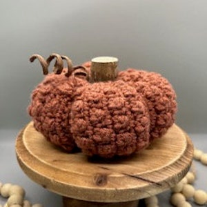 May include: A crocheted pumpkin in a terracotta colour with a brown wooden stem. The pumpkin is sitting on a wooden pedestal.
